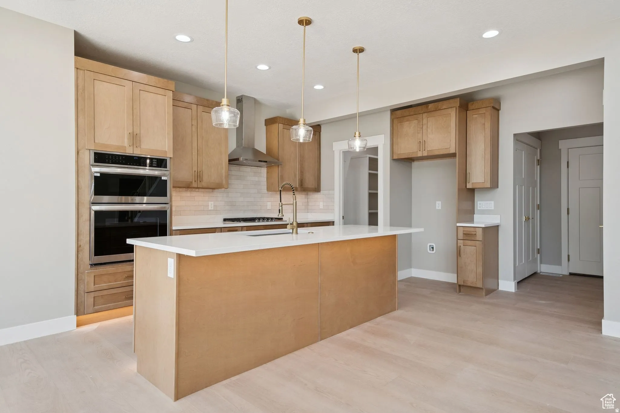 Spacious kitchen featuring light wood-finish cabinetry, a large center island with a white countertop, stainless steel double wall ovens, a coordinating range hood, and light wood-finish flooring