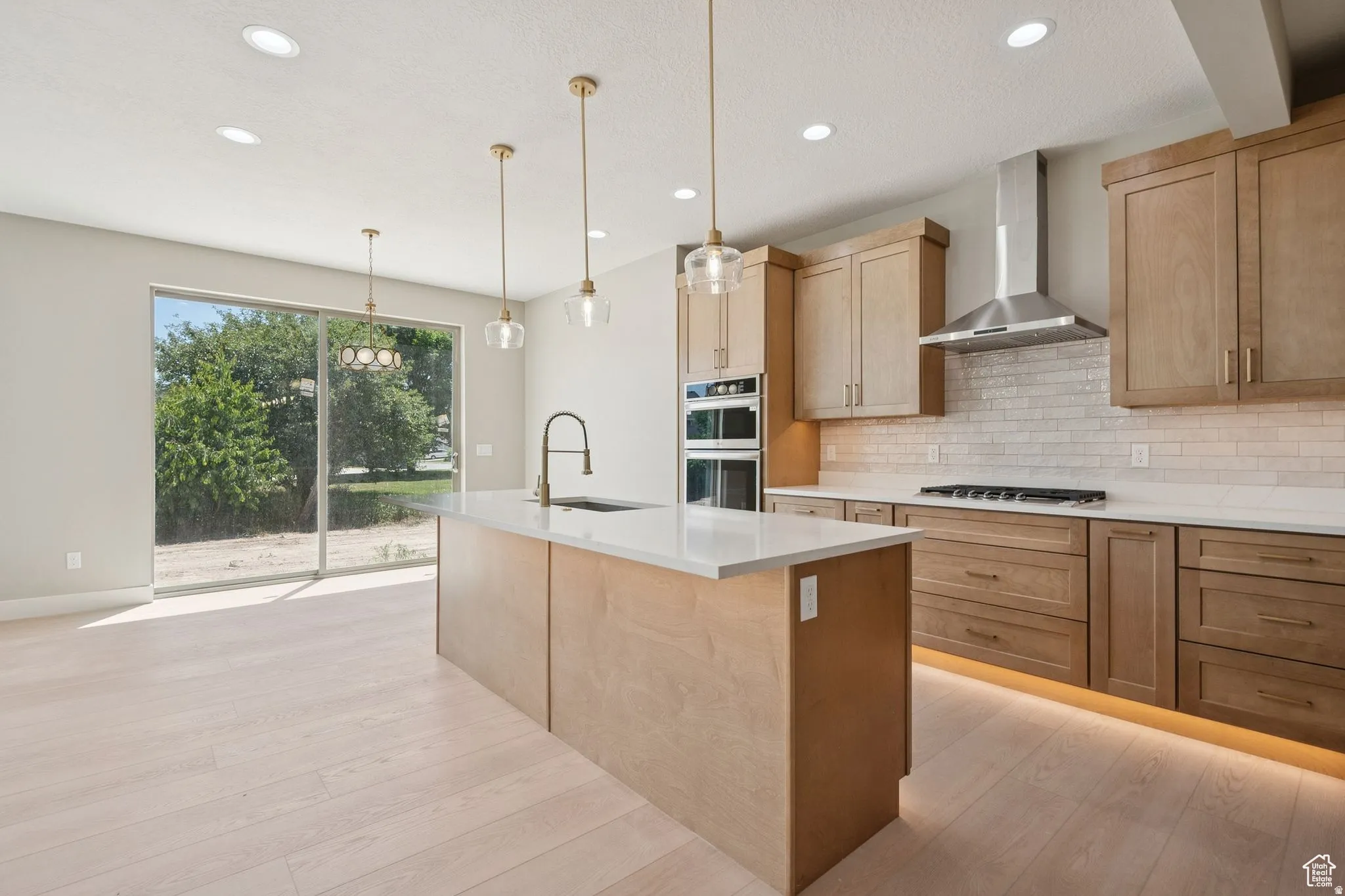 Modern kitchen featuring natural wood cabinetry, a large waterfall-edge island with wood-finish base, white quartz countertops, stainless steel appliances including a gas cooktop and wall-mounted range hood, subway tile backsplash, pendant lighting, and a