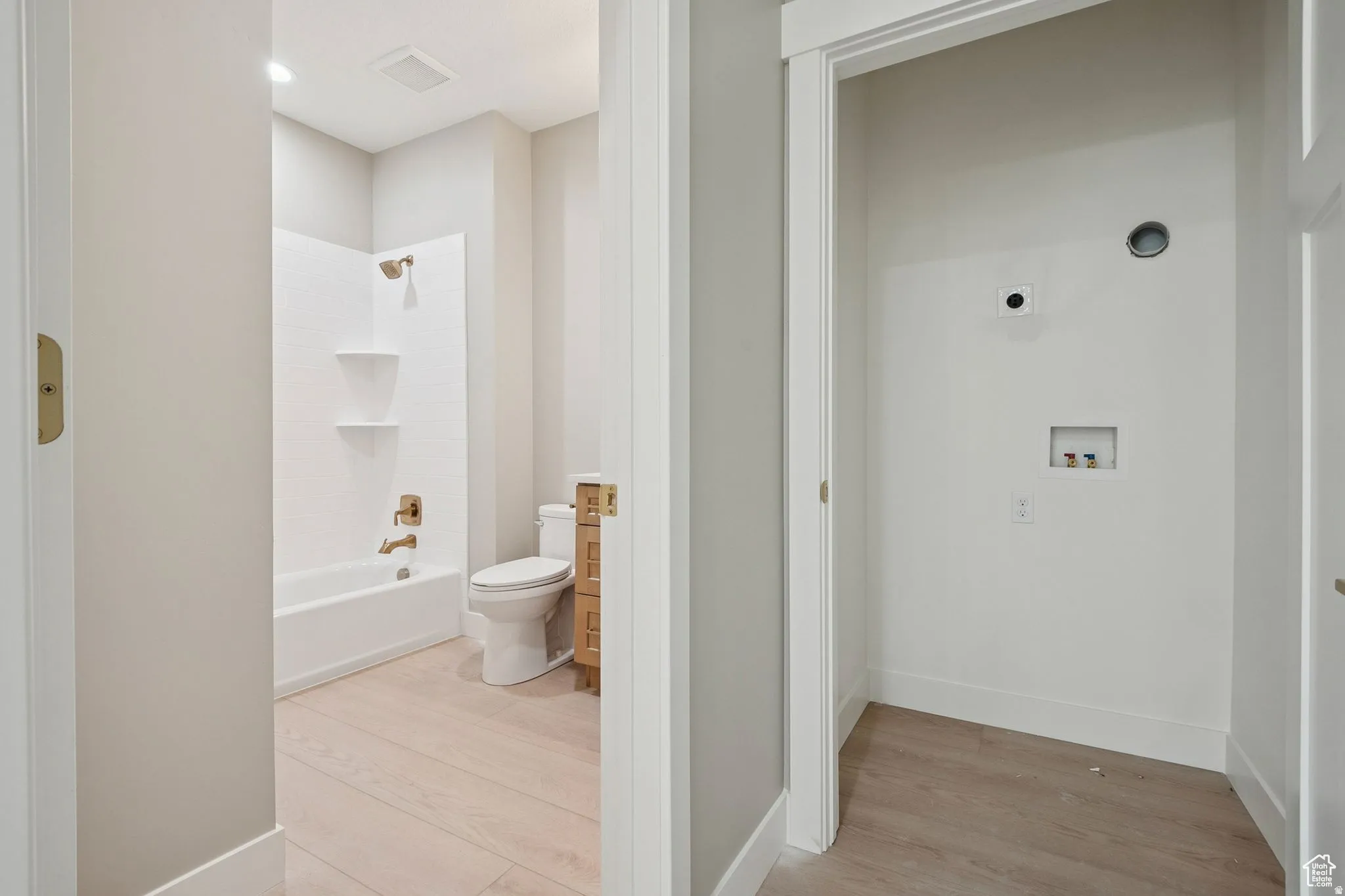 Dual-entry bathroom and laundry corridor featuring ceramic tile flooring, white subway tile shower surround with shelving, toilet, and utility space with plumbing rough-ins for future fixture installation