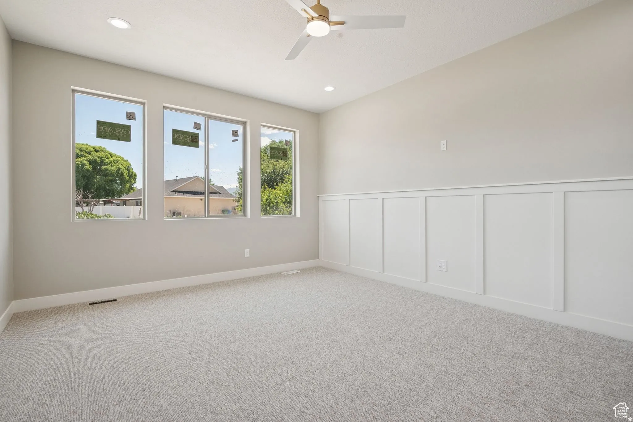 Spacious room featuring three large windows, light gray carpet flooring, white board and batten wall paneling, recessed lighting, and a ceiling fan with integrated light
