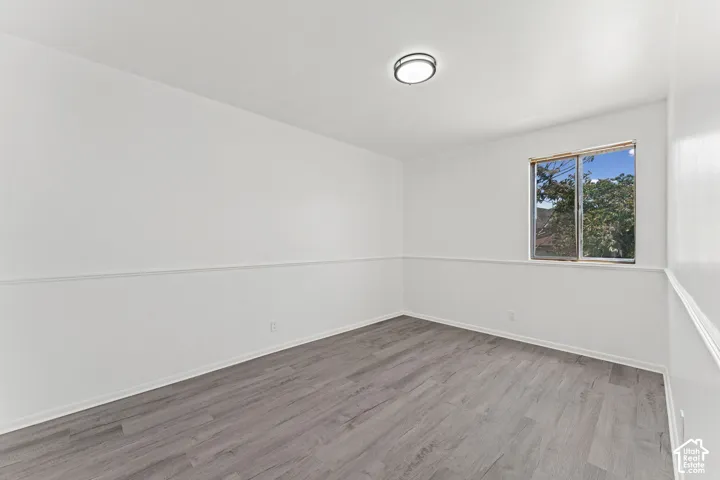 Bright bedroom featuring pristine white walls with horizontal wainscoting detail, engineered wood-finish flooring throughout, and a large double-hung window with natural light exposure and outdoor scenery