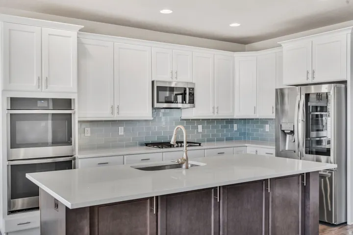 Modern kitchen featuring white shaker-style cabinetry, stainless steel appliances including a built-in microwave and double wall ovens, quartz countertops with waterfall edge on the center island, blue subway tile backsplash, and a large kitchen island wi