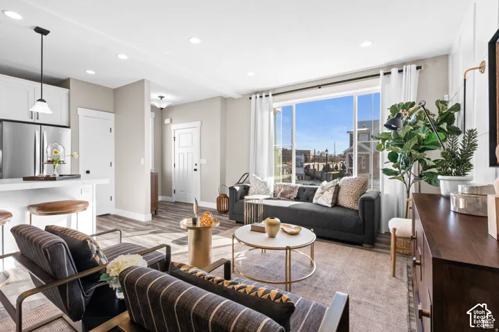 Open-concept living space showcasing floor-to-ceiling windows with cityscape backdrop, wide-plank wood-finish flooring throughout, and recessed lighting