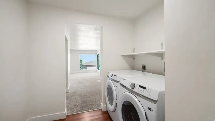 Modern laundry room featuring side-by-side front-load washers and dryers with white finish, built-in cabinetry with integrated sink, wood-finish flooring, and abundant natural light from expansive window opening to primary living spaces
