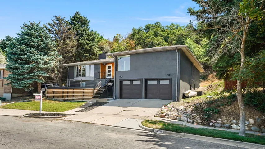 Modern split-level residence featuring a charcoal gray exterior with warm wood-tone accents, two-car attached garage with dark panel doors, and a sloped driveway with decorative stone landscaping