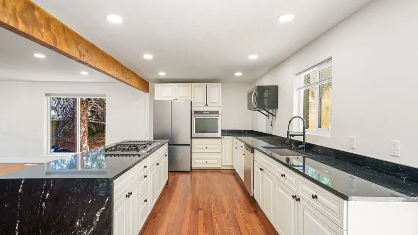 Galley kitchen featuring white cabinetry with black hardware, black quartz countertops, stainless steel appliances including a gas cooktop and built-in wall oven, undermount sink with gooseneck faucet, recessed lighting, and exposed wood beam detail