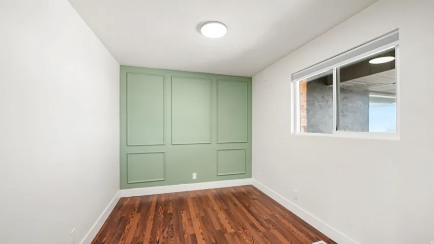 Versatile bedroom or office space featuring rich wood-finish flooring, fresh white walls, and a striking sage green accent wall with custom panel detailing