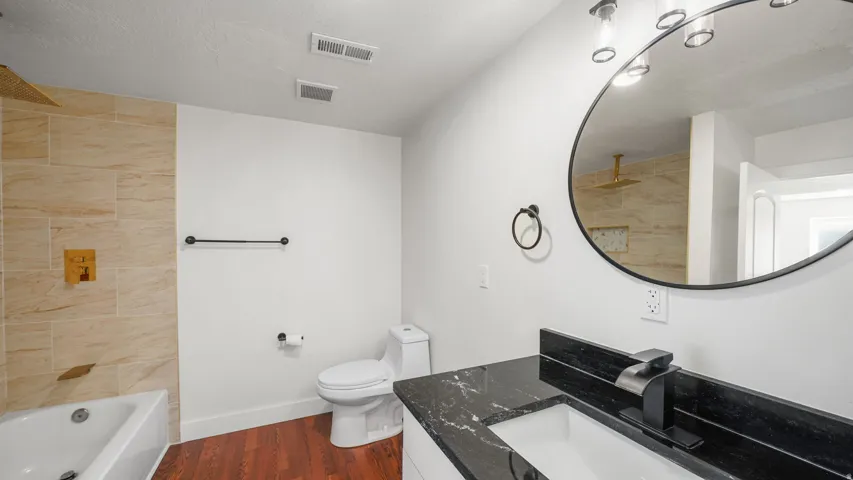 Full bathroom featuring a rectangular white bathtub with a gold-tone rain shower head, large format beige wall tiling, a toilet, and a vanity with a black countertop and integrated sink
