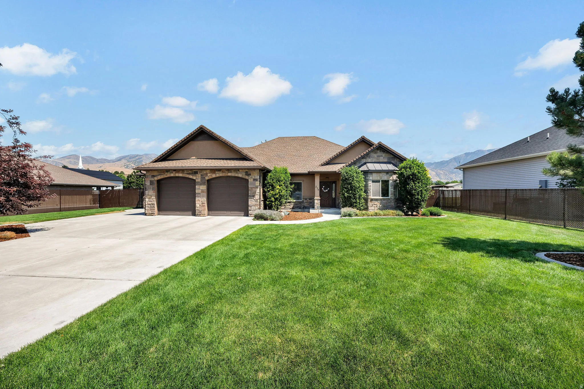 Single-story residence featuring a stone and stucco exterior, a two-car garage with arched entrances, a wide concrete driveway, and a front lawn with mature landscaping