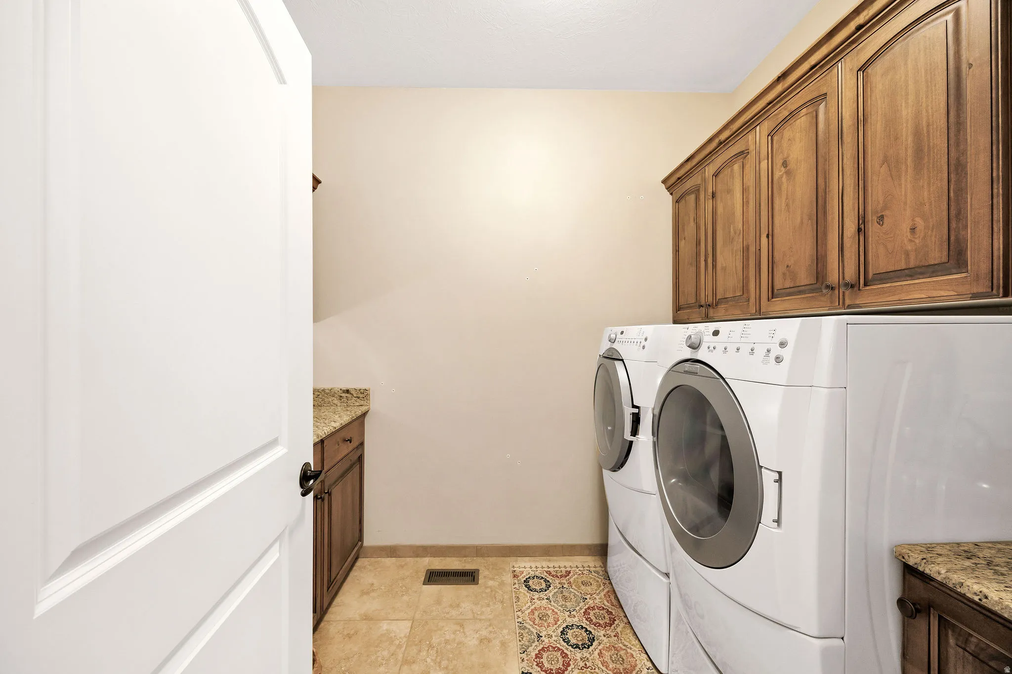 Dedicated laundry room featuring side-by-side white front-load washer and dryer, wood-finish cabinetry with ample storage, granite-look countertops, tile flooring, and recessed lighting for optimal functionality