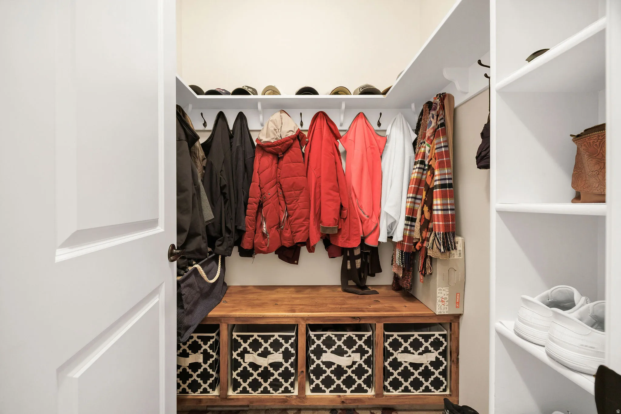 Organized entryway closet featuring built-in wooden bench with geometric patterned storage cubbies, wall-mounted rod with multiple hanging hooks, and open shelving displaying decorative baskets and accessories