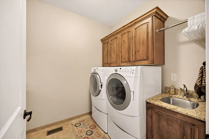 Dedicated laundry room featuring side-by-side front-load washer and dryer with white finish, custom wood cabinetry with crown molding, granite-style countertop with integrated sink, wood-finish flooring, and abundant storage