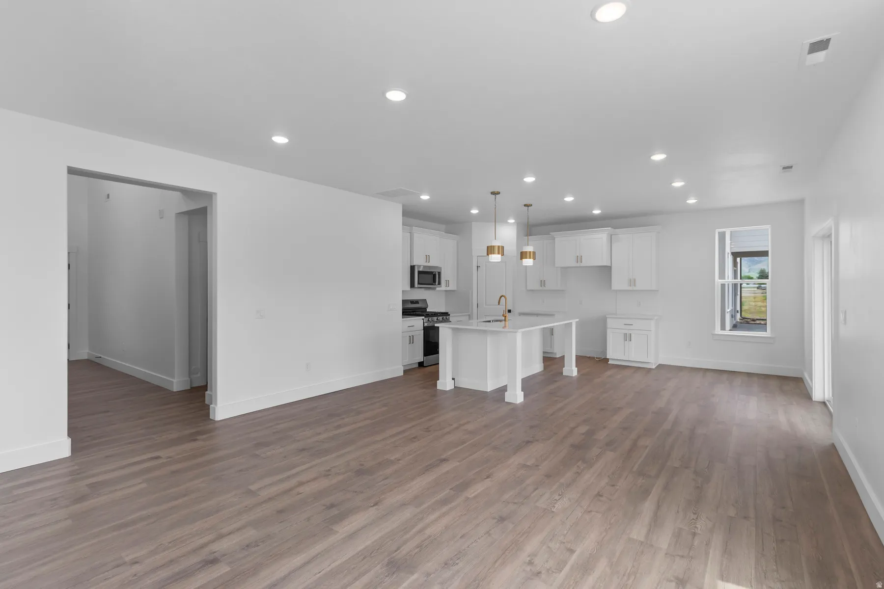 Open-concept living and kitchen area featuring recessed ceiling lighting, white cabinetry with pendant fixtures, stainless steel appliances, wood-finish flooring throughout, and abundant natural light from multiple windows and glass door access