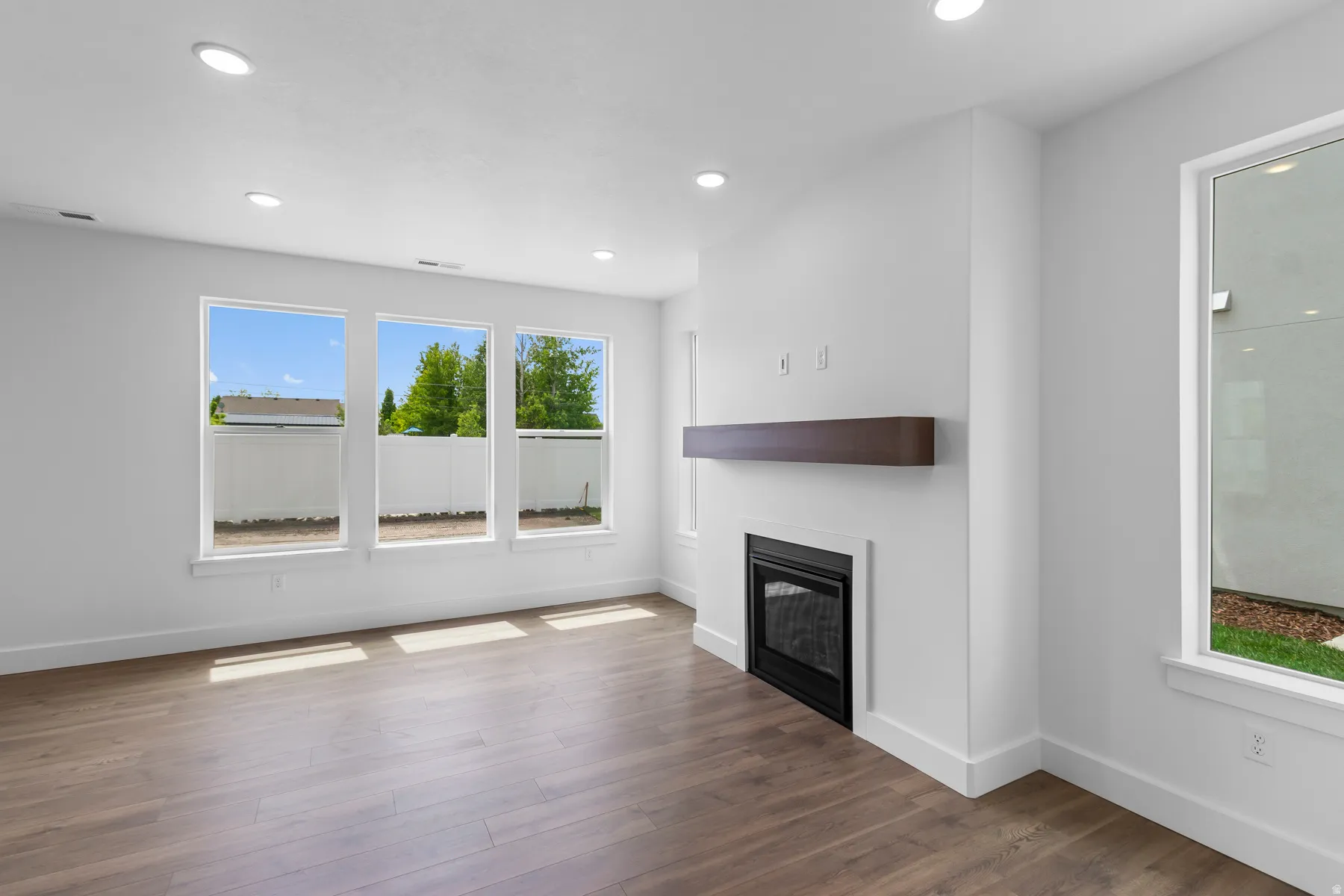 Bright, open living space showcasing gleaming wood-finish flooring, recessed ceiling lighting, and abundant natural light from triple casement windows