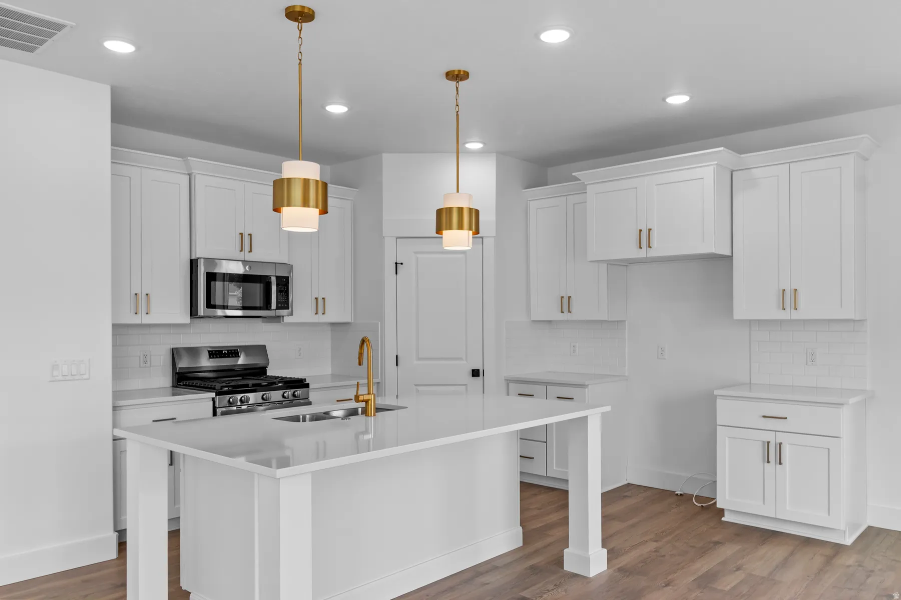 Bright, modern kitchen featuring white shaker-style cabinetry with brushed metal hardware, a large waterfall-edge quartz island with seating, stainless steel gas cooktop and built-in microwave, recessed ceiling lighting, and dual brass pendant fixtures