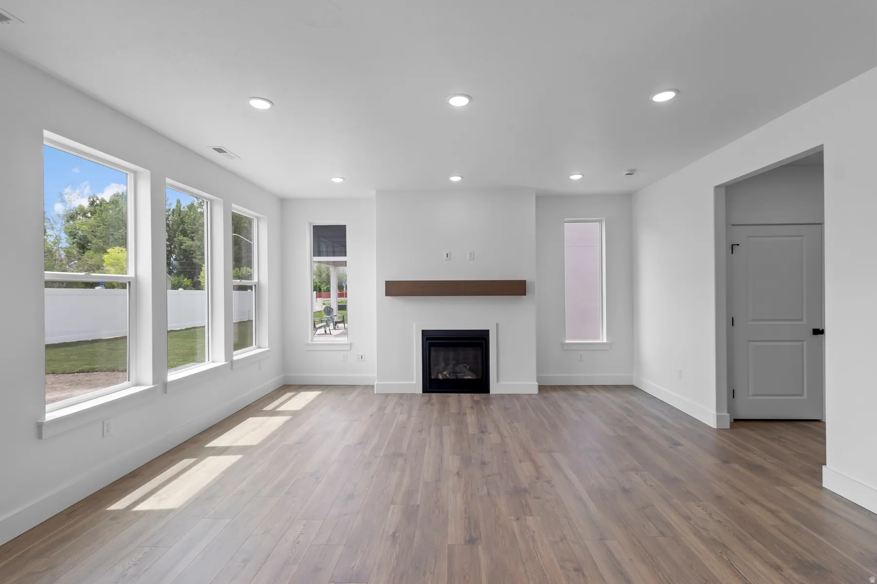 Bright and open living space showcasing gleaming wood-finish flooring, recessed ceiling lighting, and abundant natural light from expansive multi-pane windows and glass doors