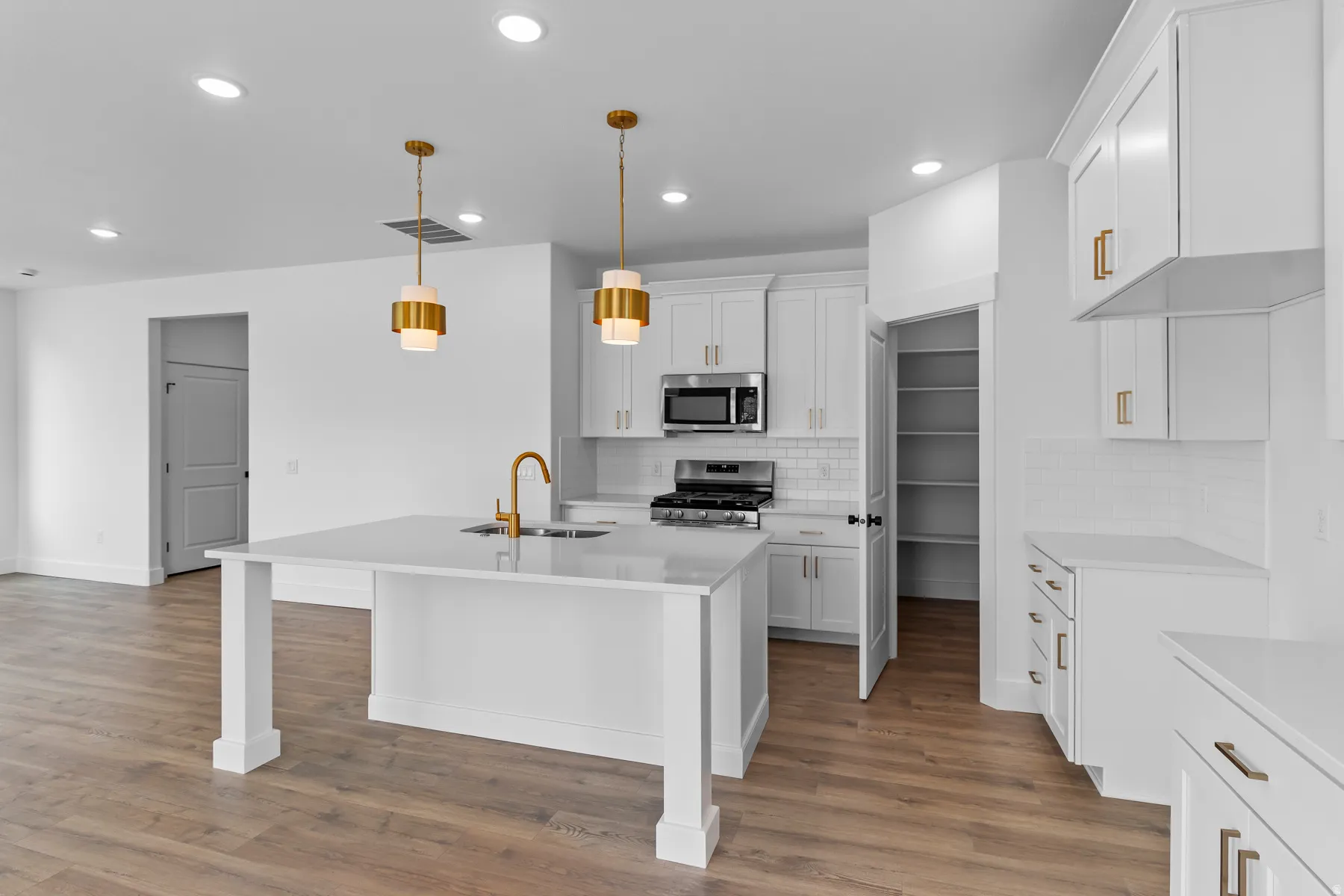 Modern kitchen featuring a large central island with an undermount sink and gold-tone faucet, white shaker-style cabinetry, stainless steel appliances, and wood-finish flooring
