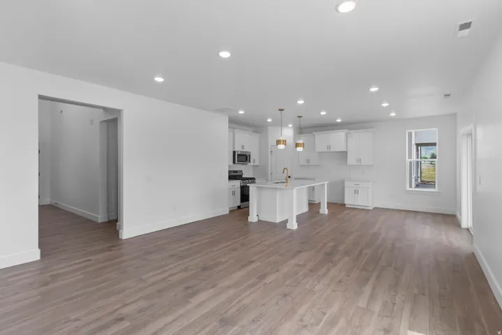 Open-concept living and kitchen area featuring recessed ceiling lighting, white cabinetry with pendant fixtures, stainless steel appliances, wood-finish flooring throughout, and abundant natural light from multiple windows and glass door access