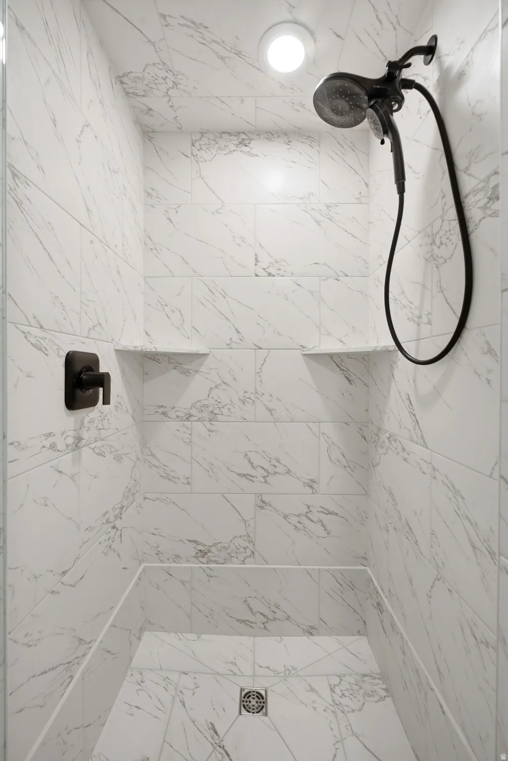 Shower stall featuring marble-look tile, matte black fixtures, recessed lighting, and integrated shelving
