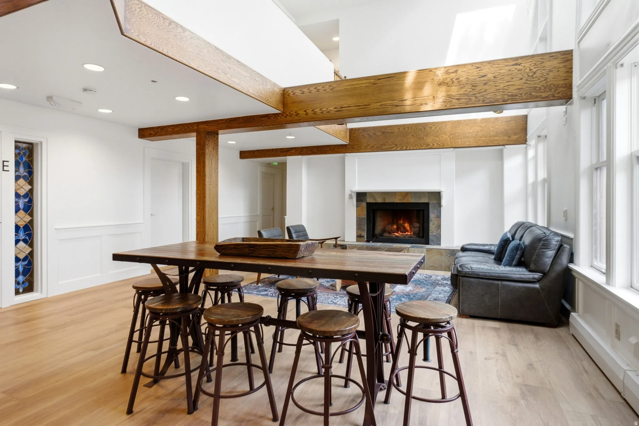 Spacious common area featuring light wood-finish flooring, exposed wood beams, a stone-surround fireplace, and multiple tall windows