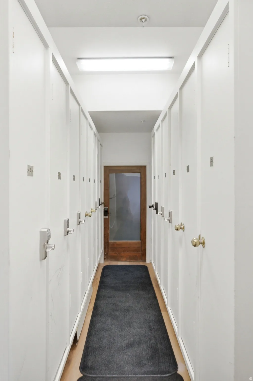 Hallway featuring multiple white storage lockers with various door handles, recessed lighting, and a wood-finish floor