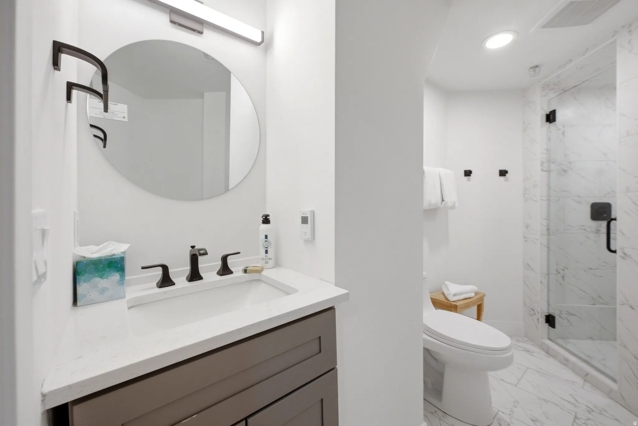 Modern bathroom featuring a vanity with a white countertop and dark gray cabinetry, a frameless oval mirror, and a walk-in shower with clear glass enclosure and marble-look tile
