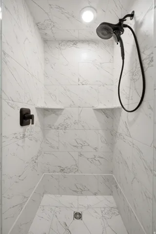 Shower stall featuring marble-look tile, matte black fixtures, recessed lighting, and integrated shelving
