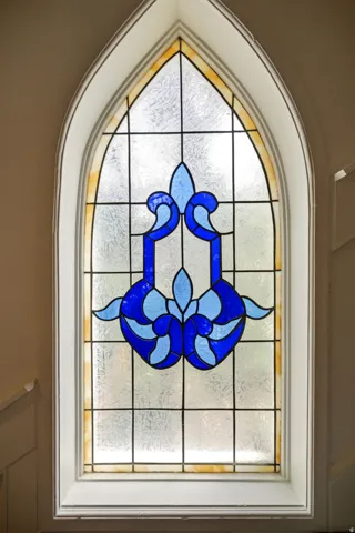 Arched stained glass window featuring blue and light blue design elements, textured glass panes, white trim, and a decorative border
