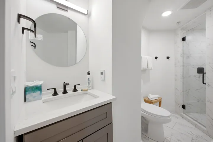 Modern bathroom featuring a vanity with a white countertop and dark gray cabinetry, a frameless oval mirror, and a walk-in shower with clear glass enclosure and marble-look tile