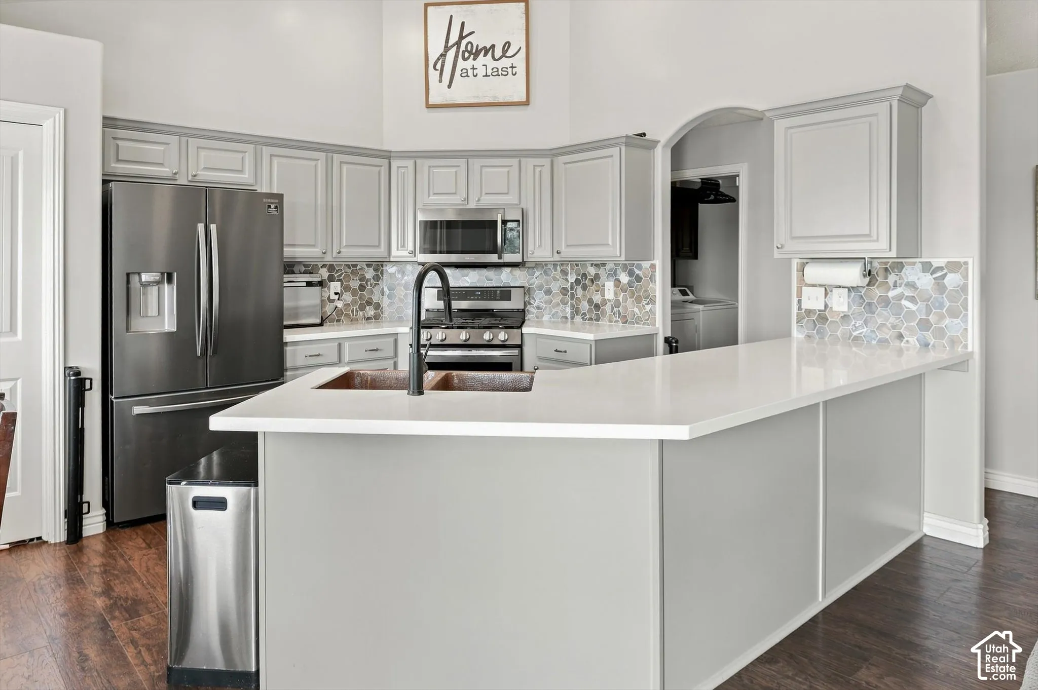 Stunning chef's kitchen featuring light gray cabinetry, stainless steel appliances including a French-door refrigerator and gas range, solid surface countertop,  mosaic tile backsplash, bar