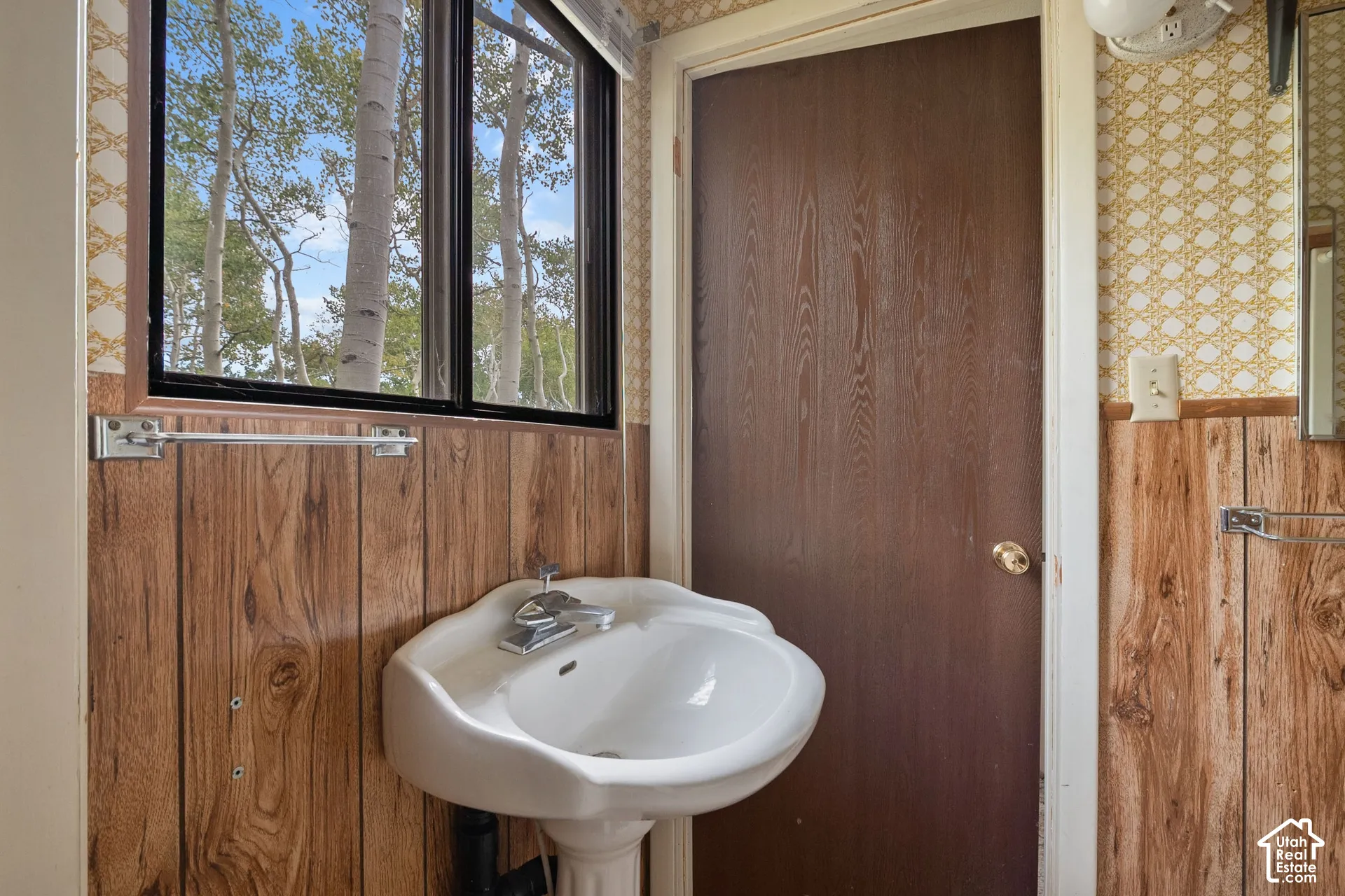 Pedestal sink with chrome fixtures, set against wood-finish paneling and patterned wallpaper