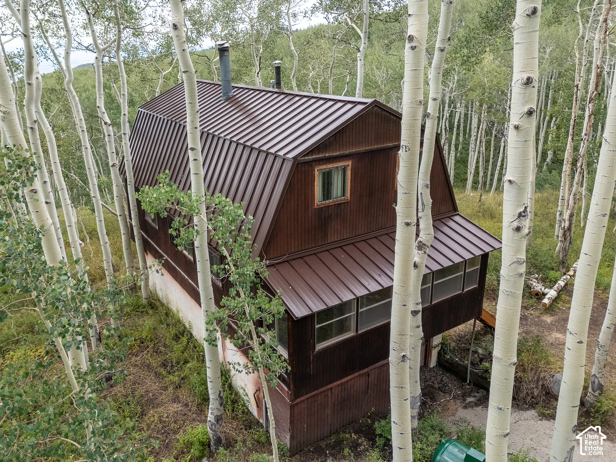 Gambrel roofline property featuring a brown metal roof, exterior wood siding, a covered porch, and mature surrounding trees
