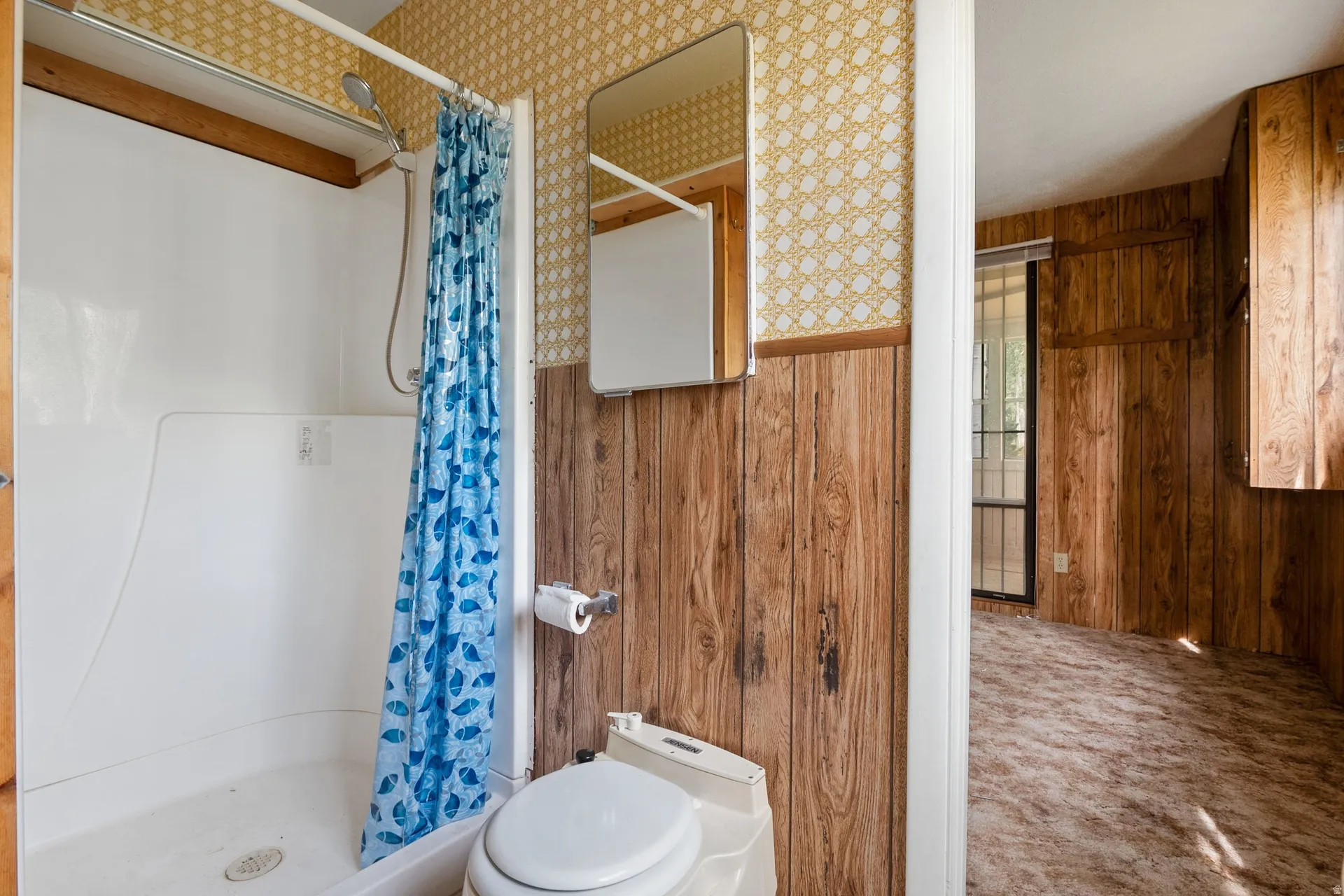 Bathroom featuring a white shower stall with an attached showerhead, RV style porta potty toilet, wood paneling, and patterned wallpaper