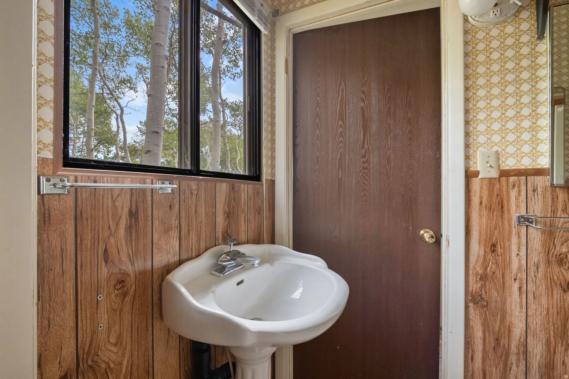 Pedestal sink with chrome fixtures, set against wood-finish paneling and patterned wallpaper