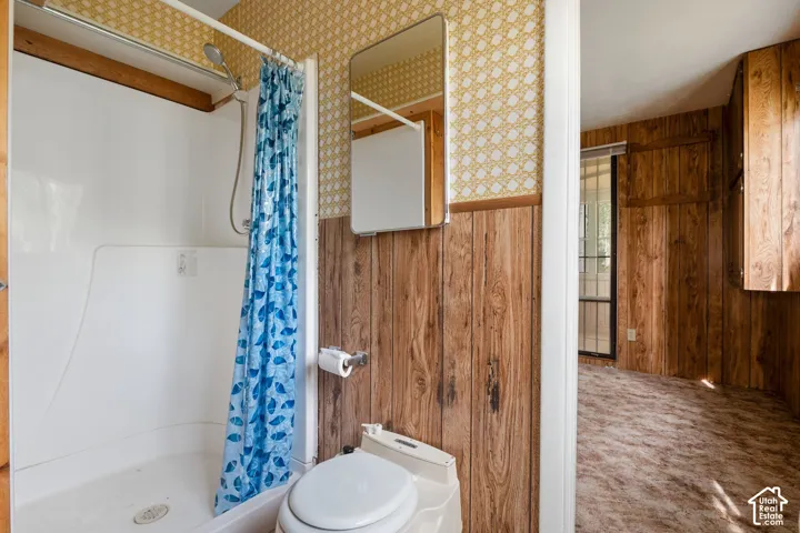 Bathroom featuring a white shower stall with an attached showerhead, RV style porta potty toilet, wood paneling, and patterned wallpaper