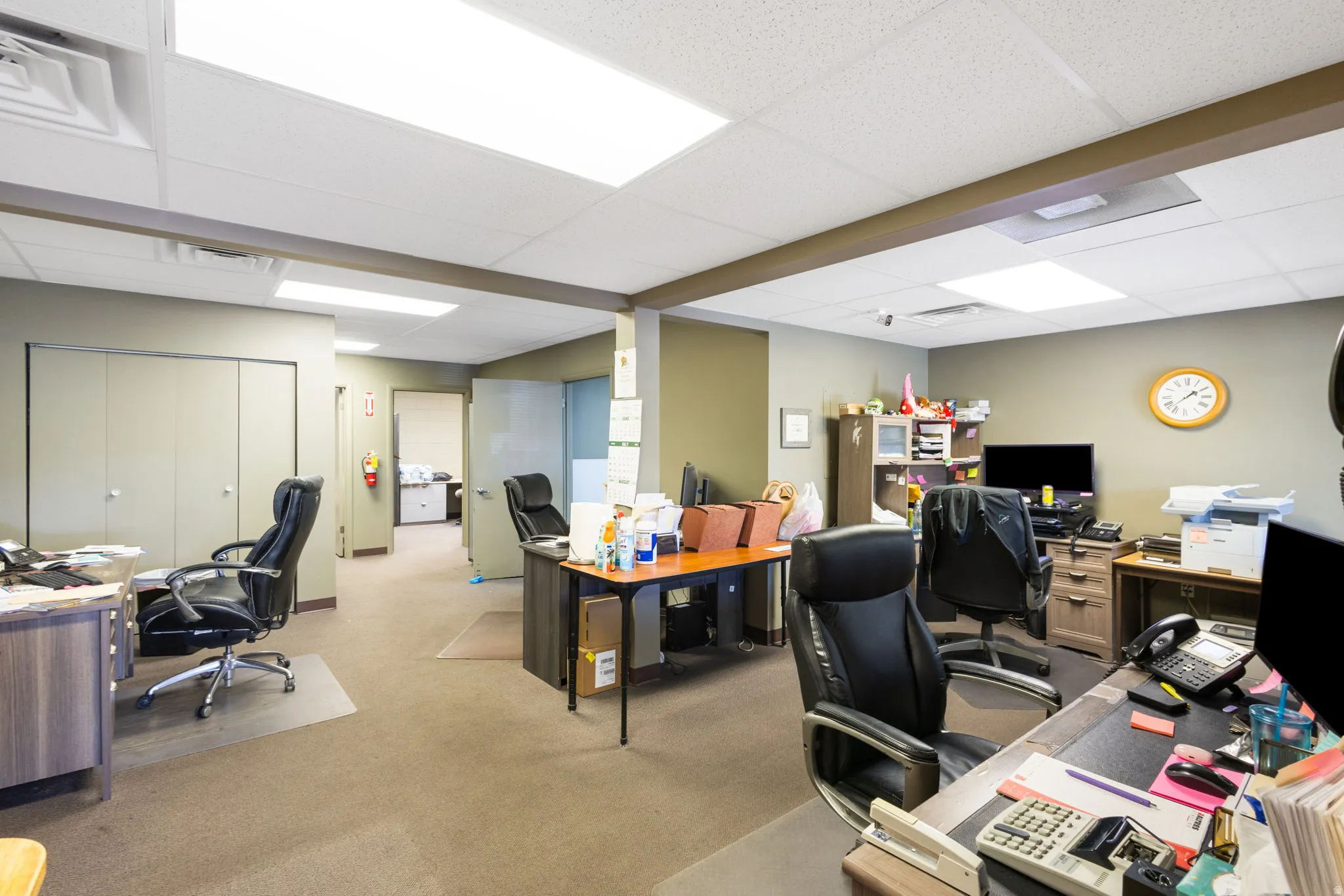 Spacious open-concept office suite featuring neutral-toned carpet flooring, recessed ceiling lighting, and multiple workstations with built-in desk surfaces