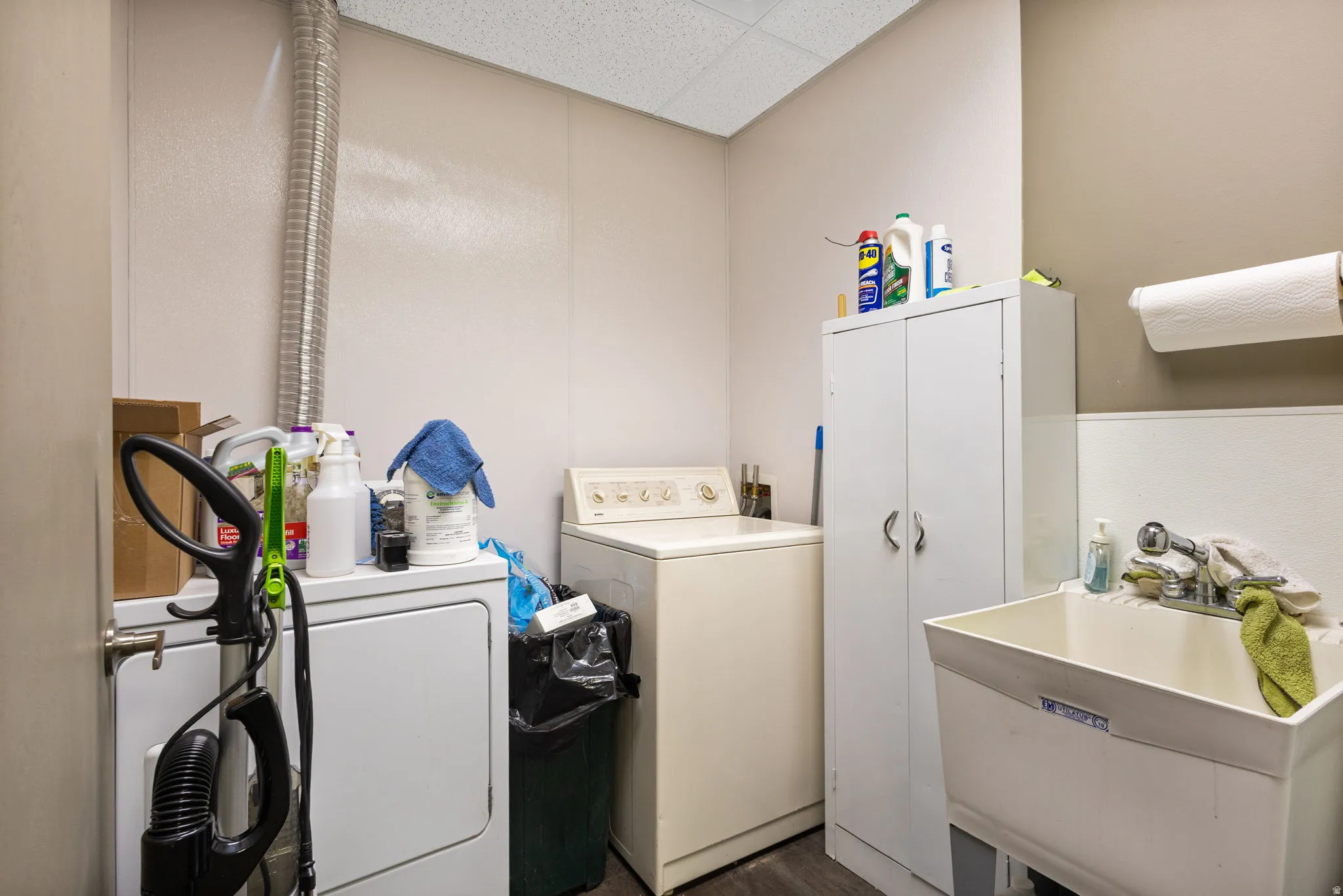 Utility room featuring full-size front-load washer and dryer, white cabinetry with ample storage, stainless steel sink with single-handle faucet, drop ceiling with recessed lighting, and organized shelving for laundry essentials and household supplies