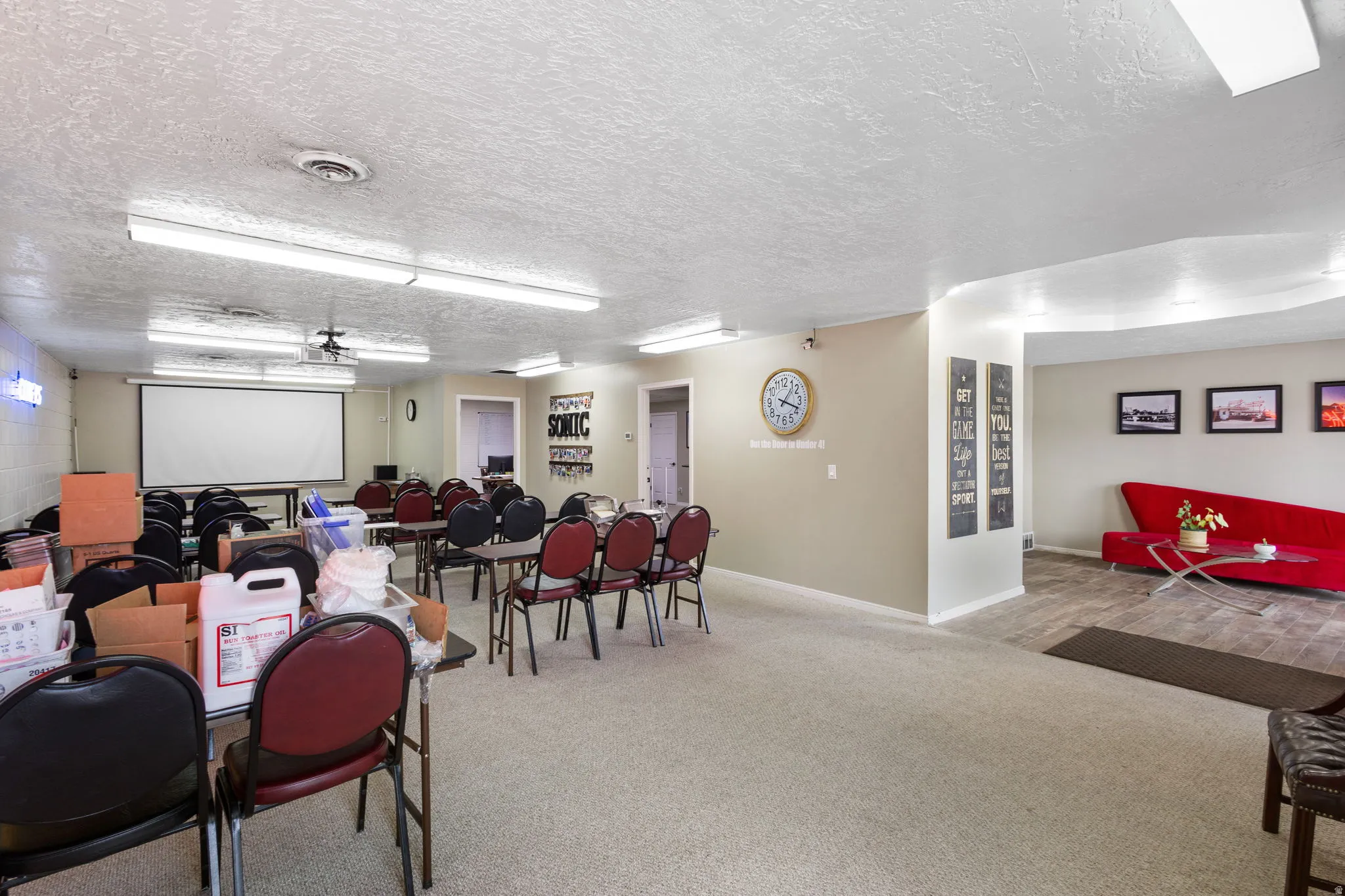 Versatile multipurpose room featuring fluorescent ceiling lighting, a wall-mounted projector screen, carpeted flooring, and flexible seating arrangements