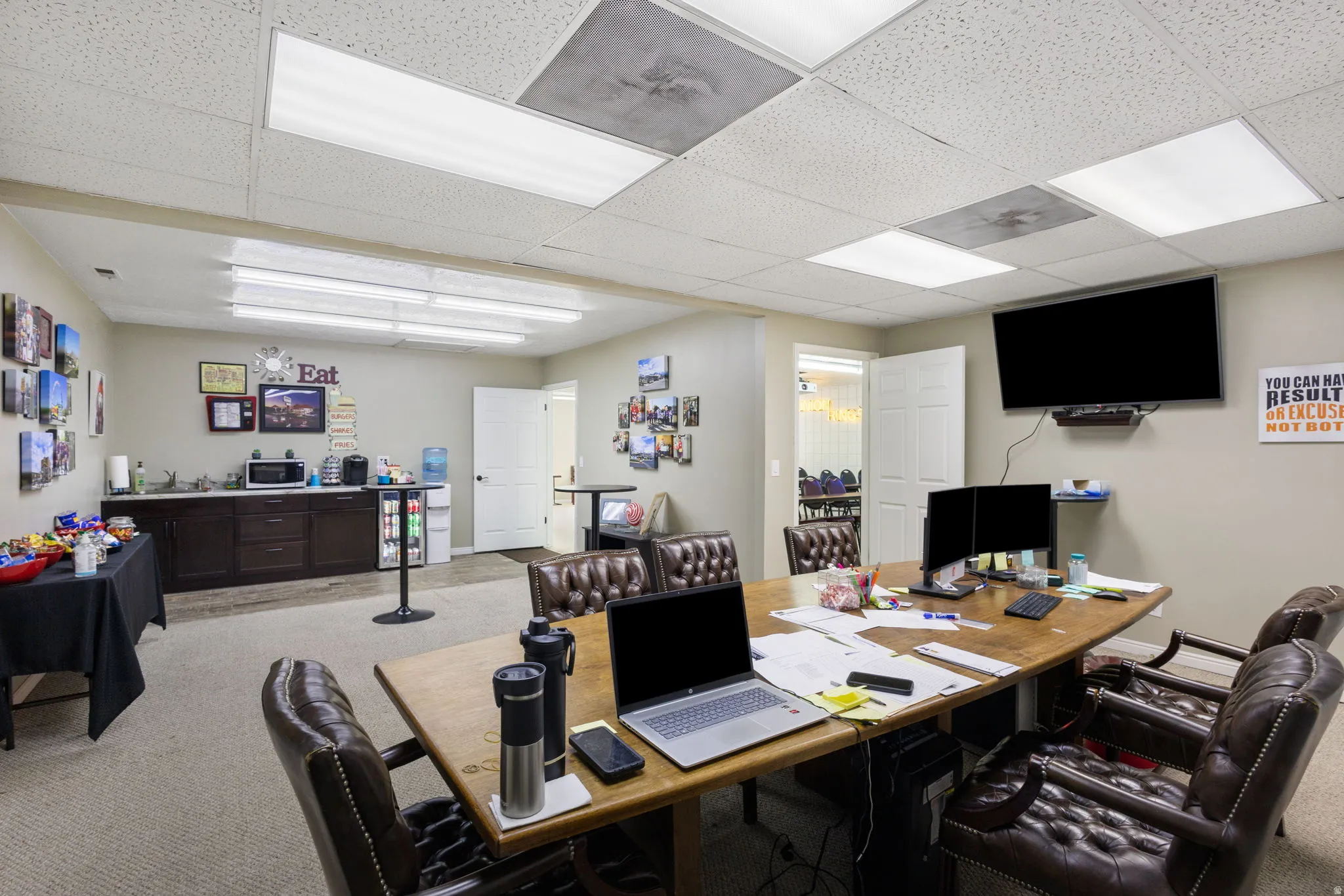 Professional office space featuring a spacious open layout with recessed and panel lighting throughout, multiple work stations with wood-finish desks, ergonomic seating, wall-mounted flat-screen display, built-in dark cabinetry with storage, and carpeted