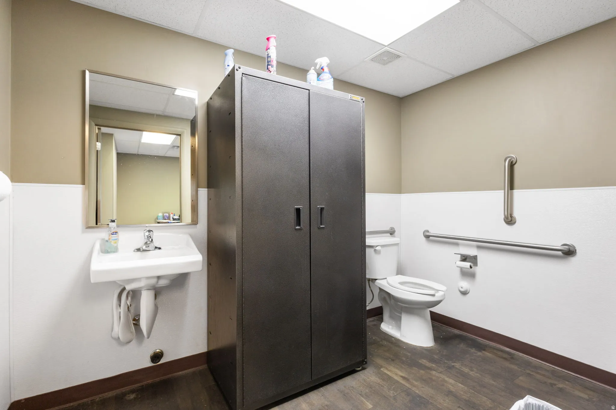 Restroom featuring wood-finish flooring, a wall-mounted sink with mirror, a commode, and polished chrome grab bars