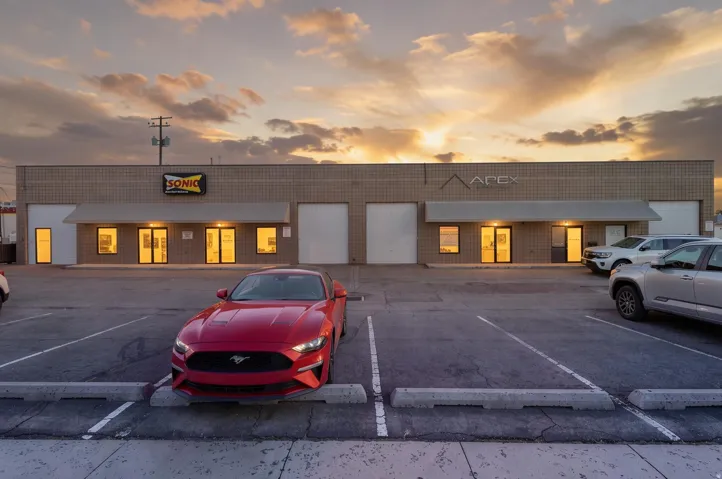 Modern commercial property featuring a spacious asphalt parking lot with clearly marked spaces, metal-frame warehouse structure with illuminated storefront windows, and multiple garage bays with white roll-up doors