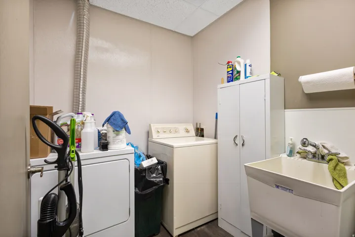 Utility room featuring full-size front-load washer and dryer, white cabinetry with ample storage, stainless steel sink with single-handle faucet, drop ceiling with recessed lighting, and organized shelving for laundry essentials and household supplies