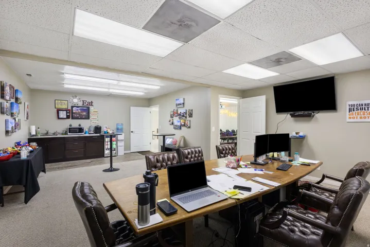 Professional office space featuring a spacious open layout with recessed and panel lighting throughout, multiple work stations with wood-finish desks, ergonomic seating, wall-mounted flat-screen display, built-in dark cabinetry with storage, and carpeted