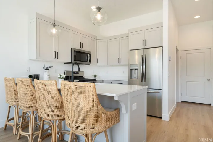 Modern kitchen featuring white shaker-style cabinetry with hardware accents, stainless steel appliances including a built-in microwave and refrigerator, a waterfall-edge white quartz island with seating for multiple guests, and wood-finish flooring throug