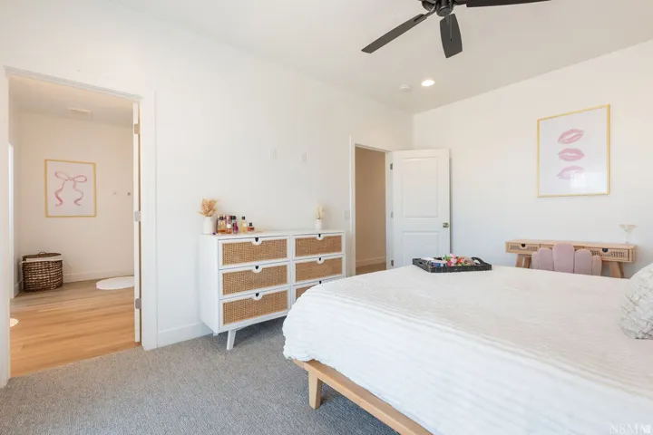 Spacious primary bedroom featuring a wood-finish platform bed, gray carpeting, and abundant natural light from recessed ceiling fixtures and ceiling fan