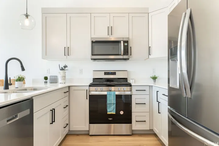 Modern kitchen featuring crisp white cabinetry with brushed metal hardware, stainless steel appliances including a gas range, microwave, and French-door refrigerator, white subway tile backsplash, light wood-finish flooring, and a curved pendant light fix