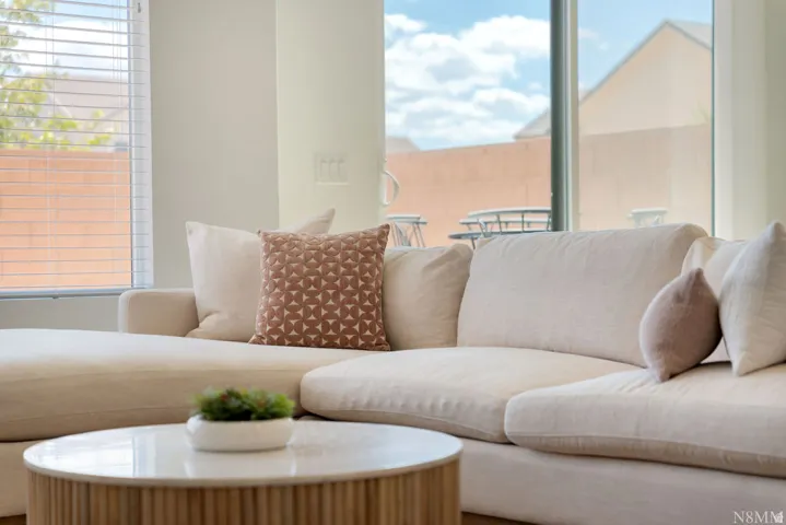Bright, light-filled living room showcasing floor-to-ceiling windows, a curved sectional sofa with neutral upholstery, a wood-finish round cocktail table, and an abundance of natural light