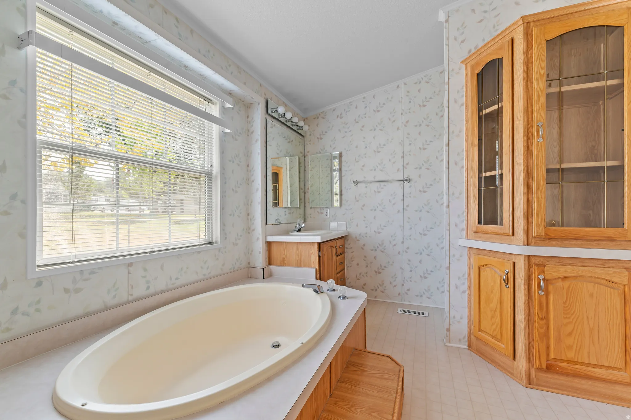 Spacious bath featuring an oval soaking tub, a double window with blinds, a vanity with a mirror and strip lighting, and a built-in wood-finish cabinet with glass inserts