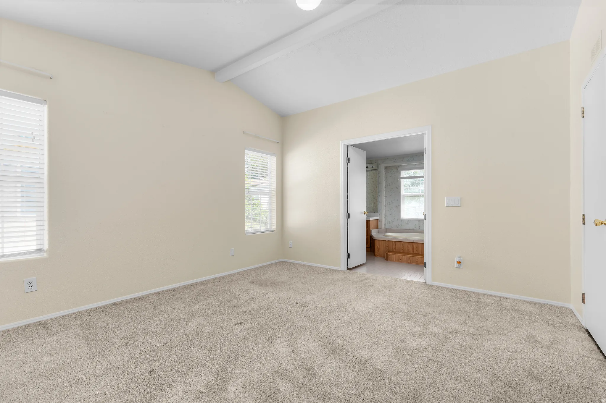 Spacious room featuring vaulted ceilings, two windows with blinds, light-colored carpeting, and a doorway opening to a bathroom with a built-in tub
