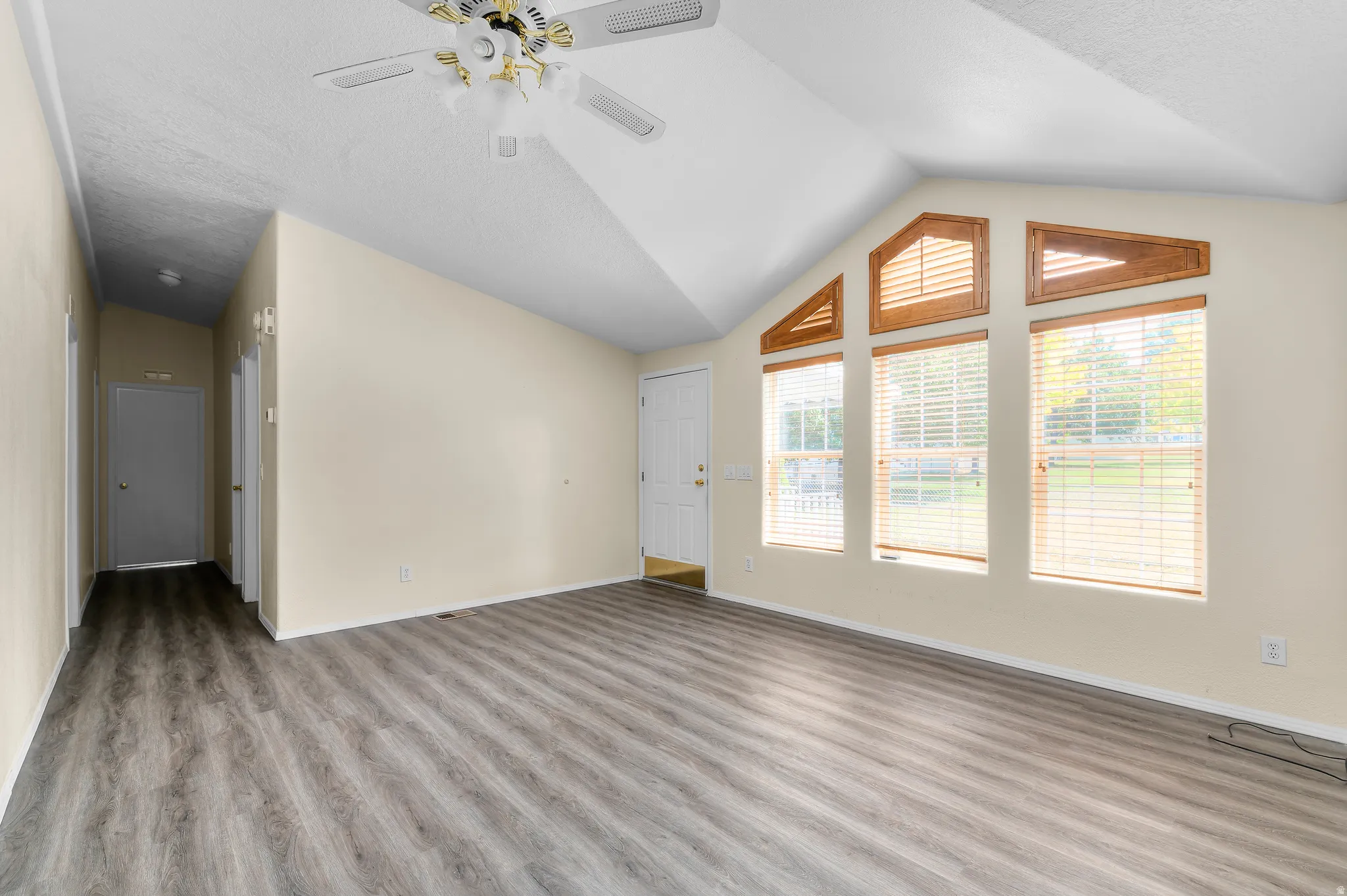 Spacious room featuring vaulted ceilings, wood-finish flooring, a ceiling fan with integrated lighting, and a series of three windows with integrated wood-frame transom windows.